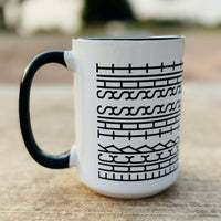 A white ceramic coffee mug with black text and geometric patterns wrapped around the surface, displayed against an outdoor background.
