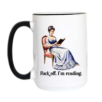 A white ceramic mug with a black handle and black rim, featuring a vintage-style illustration of a woman in a blue gown reading a book, with the text 'Fuck off. I'm reading.