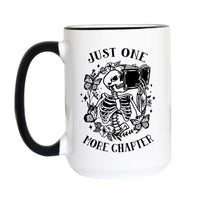 A white ceramic mug with a black handle and black rim, featuring a black graphic of a skeleton holding an open book surrounded by flowers, with the text 'JUST ONE MORE CHAPTER'.