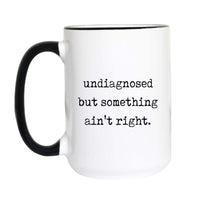 Funny meme coffee mug with black typewriter text reading "undiagnosed but something ain't right." on a white mug with black accent handle and rim