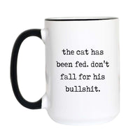 Funny cat lover coffee mug with black typewriter text reading "the cat has been fed. don't fall for his bullshit." on a white mug with black accent handle and rim