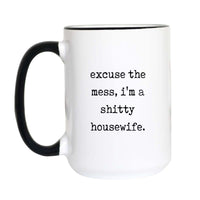 Funny housewife coffee mug with black typewriter text reading "excuse the mess, i'm a shitty housewife" on a white mug with black accent handle and rim