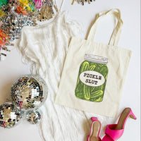Tote bag with pickle design and text on a white surface with disco balls and high heels.