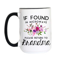 A white ceramic mug with a black handle and black rim, featuring a humorous printed message that reads 'IF FOUND IN MICROWAVE PLEASE RETURN TO Grandma' in black text, with a pink and purple floral illustration in the center.