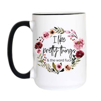 A white ceramic mug with a black handle and black rim, featuring a floral wreath graphic and the text 'I like pretty things & the word fuck' on the front.
