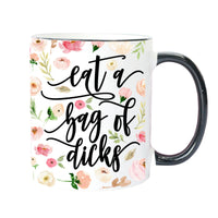 A white ceramic mug with a black handle and black rim, decorated with a watercolor floral pattern and the phrase 'eat a bag of dicks' in black script.