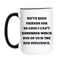 A white ceramic mug with a black handle and black rim, featuring the humorous text 'WE'VE BEEN FRIENDS FOR SO LONG I CAN'T REMEMBER WHICH ONE OF US IS THE BAD INFLUENCE.' printed in bold black capital letters on the front.