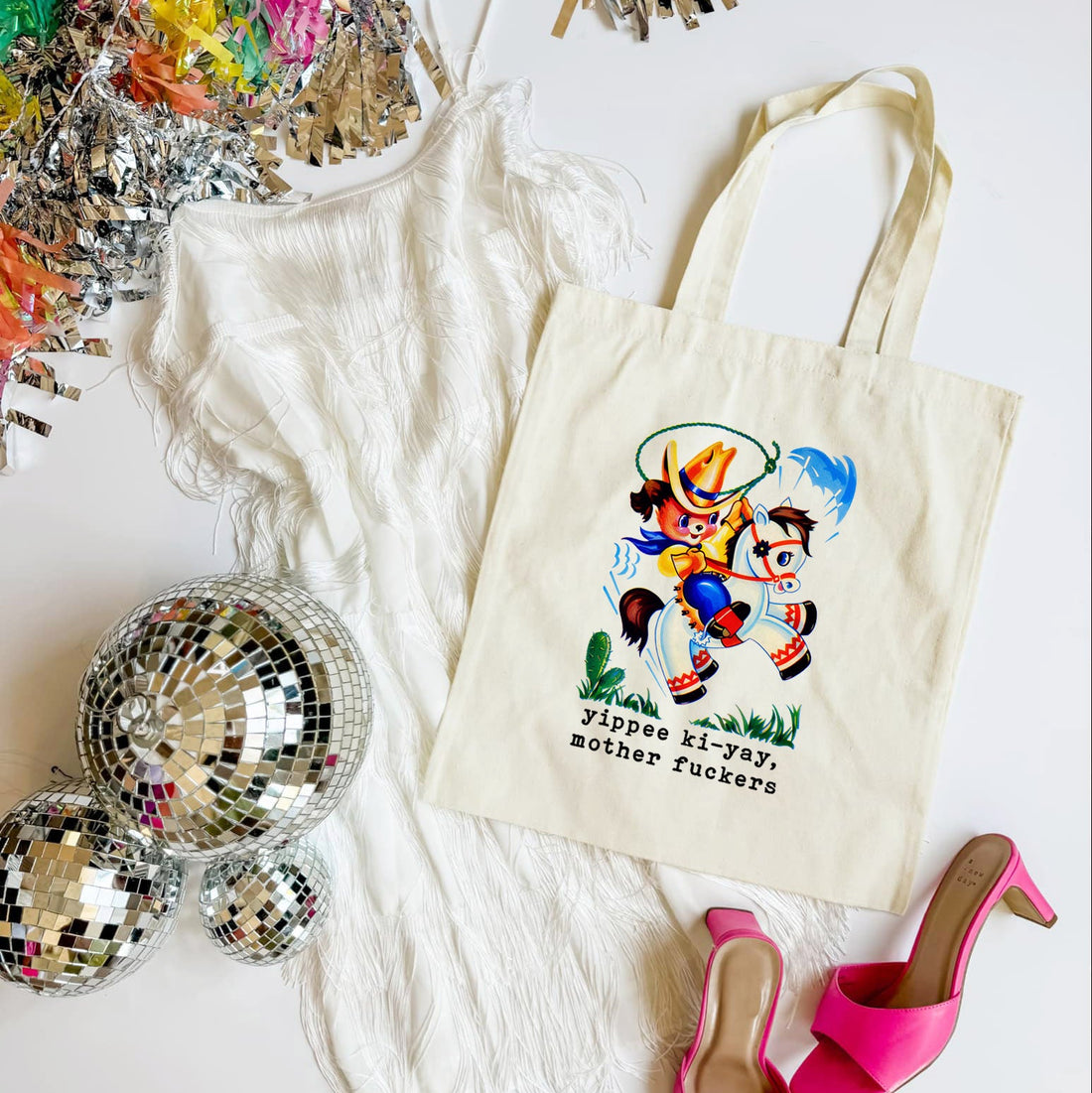 Tote bag with cartoon character and text, surrounded by disco balls and high heels on a white surface