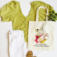 Green top, white pants, beige tote bag with cat illustration, and brown sandals on a white background