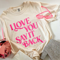 Beige t-shirt with pink text 'I LOVE YOU SAY IT BACK' on a white surface with pink sunglasses and decorative items.