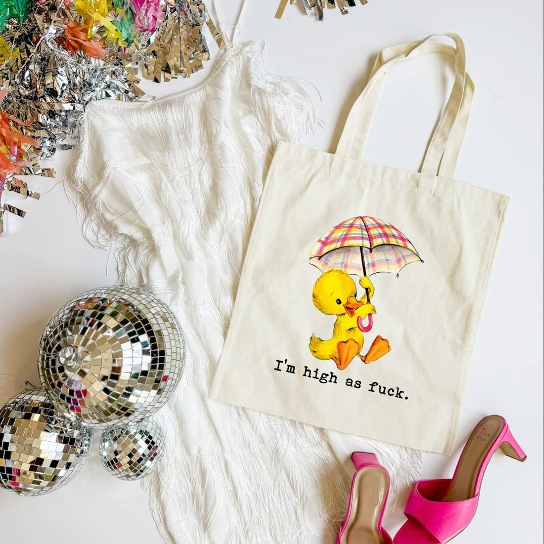 Tote bag with a duck holding an umbrella design and text, surrounded by disco balls and high heels on a white surface.