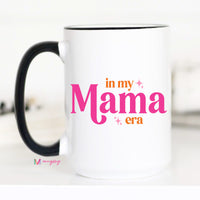 White mug with black handle featuring 'in my Mama era' text in pink and orange on a white background.