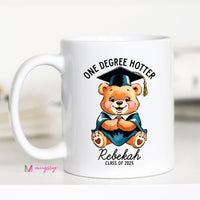 A white ceramic coffee mug with a black handle featuring a cartoon bear wearing a graduation cap and the text 'One Degree Hotter', with a personalized name 'Rebekah' and 'Class of 2021'.