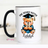 A white ceramic coffee mug with a black handle featuring a cartoon bear wearing a graduation cap and the text 'One Degree Hotter', with a personalized name 'Rebekah' and 'Class of 2021'.