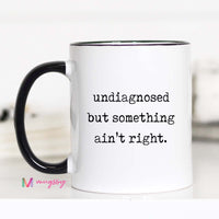 White mug with black interior and handle, featuring text 'undiagnosed but something ain't right.' on a white background.