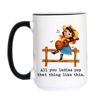 A white ceramic mug with a black handle and black rim, featuring a cartoon illustration of a girl playing a guitar on a fence with the text 'All you ladies pop that thing like this.'.