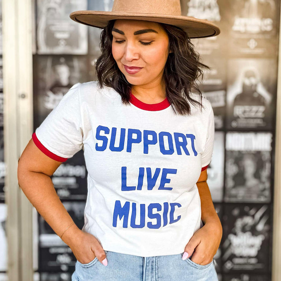 Woman wearing a white t-shirt with 'Support Live Music' text, standing in front of a store entrance.