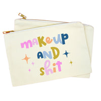 An off-white canvas zippered pouch with gold-tone zippers and a colorful retro-style text graphic reading 'makeup and shit' in pink, blue, yellow, and orange, with small star accents, shown on a plain white background.