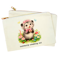 An off-white canvas zipper pouch featuring a cute illustration of a hedgehog wearing a pink bonnet and bow, surrounded by colorful flowers, with the text 'currently crashing out' printed below. A second pouch is partially visible behind it.