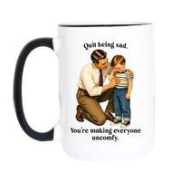 A white ceramic mug with a black handle and black rim, featuring a printed illustration of a man comforting a young boy with the text 'Quit being sad. You're making everyone uncomfy.