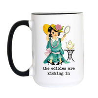 A white ceramic coffee mug with a black handle and black rim, featuring a retro illustration of a girl in a green dress and yellow hat looking in a mirror, with the text 'the edibles are kicking in' printed below.