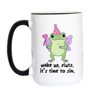 A white ceramic mug with a black handle and black rim, featuring a cartoon green frog wearing a pink party hat and wand, with the text 'wake up, sluts. it's time to sin.
