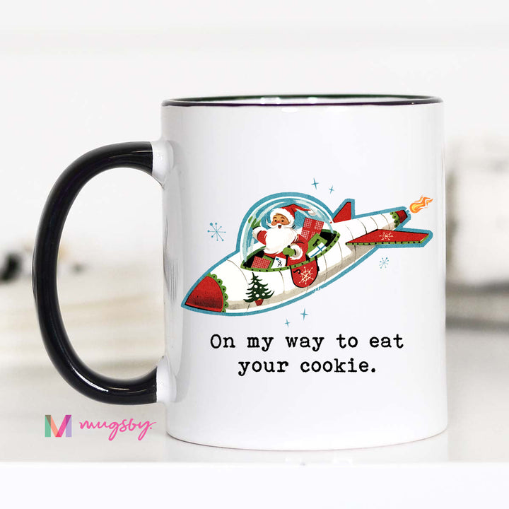 A white ceramic mug with a colorful illustration of Santa Claus riding a rocket, surrounded by snowflakes and a Christmas tree, and the text 'On My Way To Eat Your Cookie'.