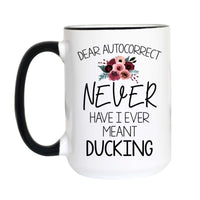 A white ceramic mug with a black handle and black rim, featuring the humorous text 'DEAR AUTOCORRECT NEVER HAVE I EVER MEANT DUCKING' in black lettering, accented by a small watercolor floral design in pink, burgundy, and green.