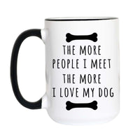A white ceramic mug with a black handle and black interior rim, printed with the phrase 'THE MORE PEOPLE I MEET THE MORE I LOVE MY DOG' between two black bone illustrations.