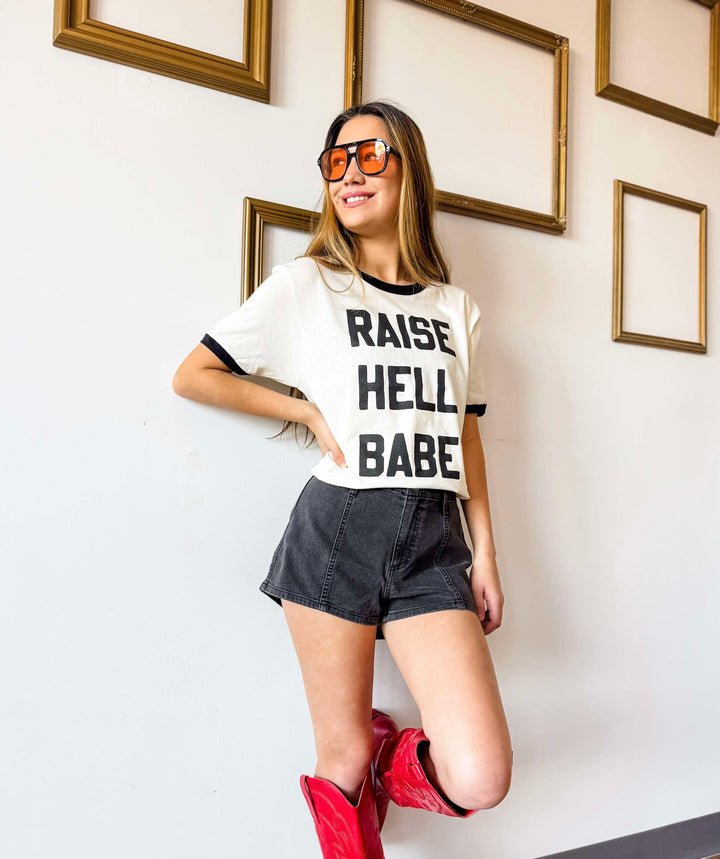 Woman wearing a t-shirt with 'Raise Hell Babe' text, denim shorts, and red boots in a room with gold-framed pictures.