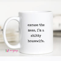 White mug with text 'excuse the mess, i'm a shitty housewife.' on a blurred background