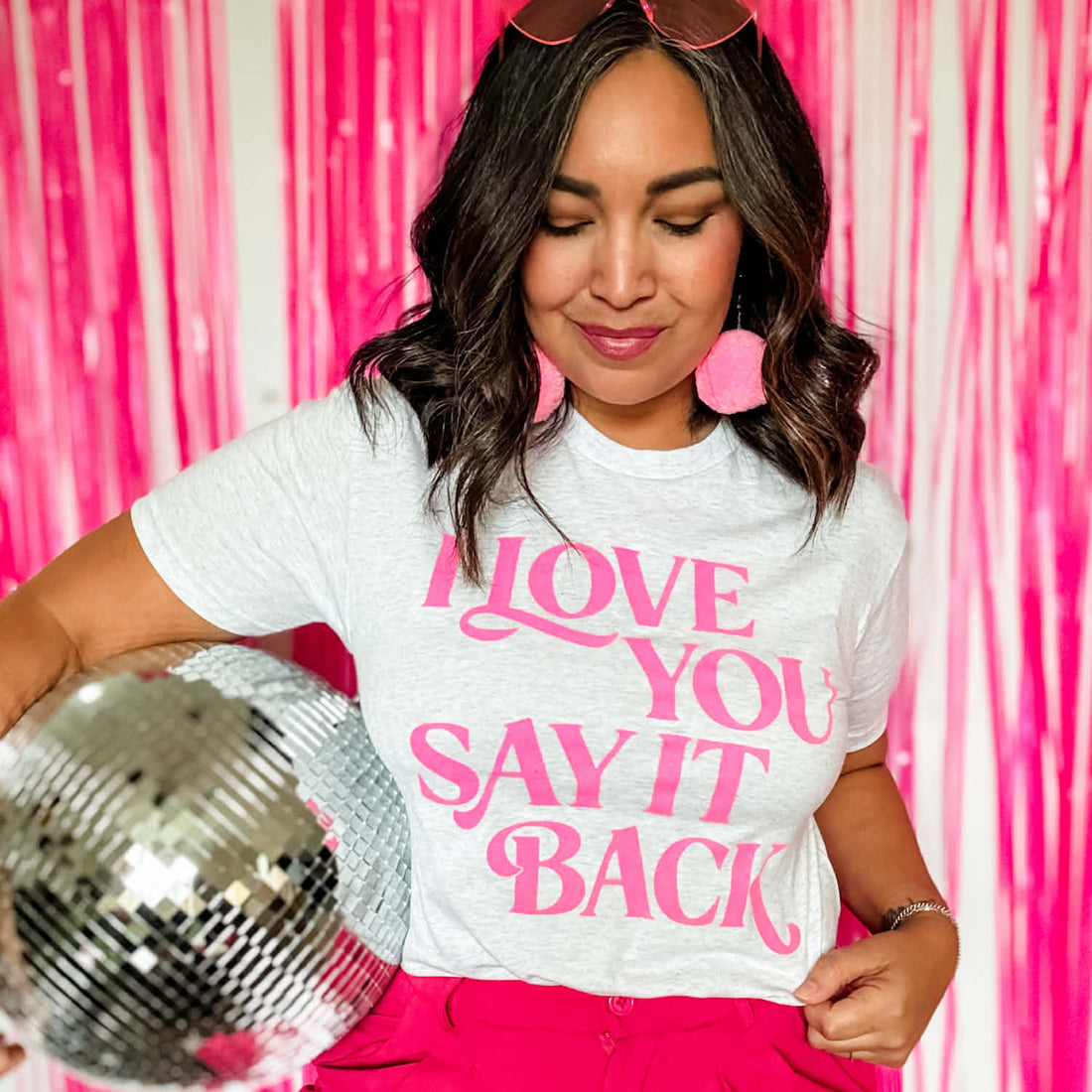 Woman wearing a white t-shirt with pink text, holding a disco ball in front of a pink curtain.
