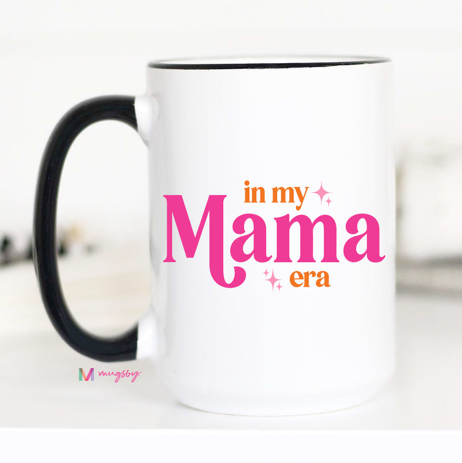 White mug with black handle featuring 'in my Mama era' text in pink and orange on a white background.