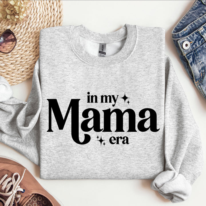 Gray sweatshirt with 'in my Mama era' text on a white surface with accessories.