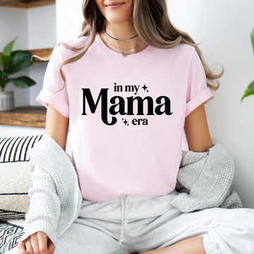 Person wearing a pink t-shirt with 'in my Mama era' text, sitting indoors.