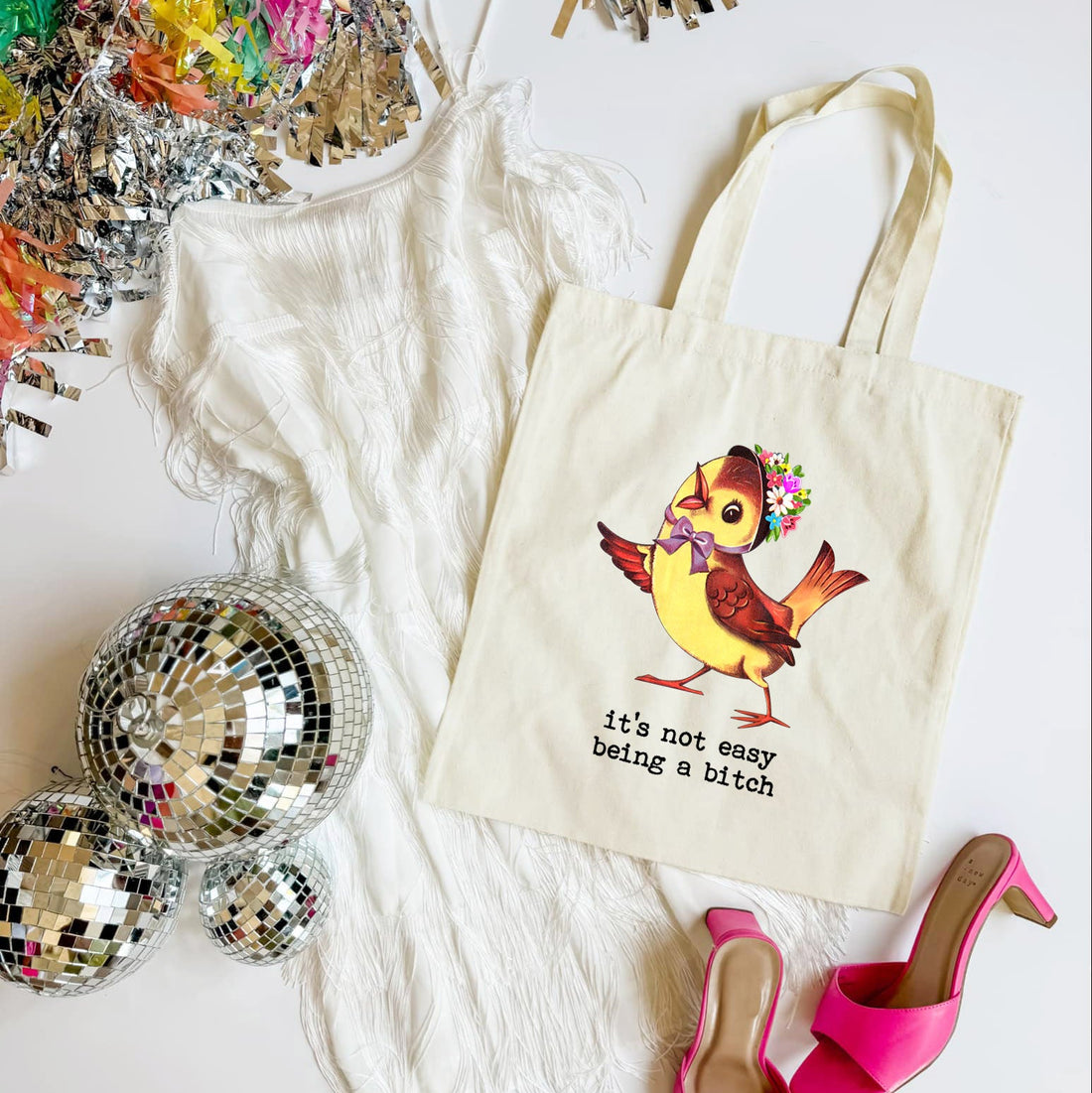 Tote bag with a colorful bird design and text, surrounded by disco balls and high heels on a white surface.