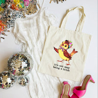 Tote bag with a colorful bird design and text, surrounded by disco balls and high heels on a white surface.
