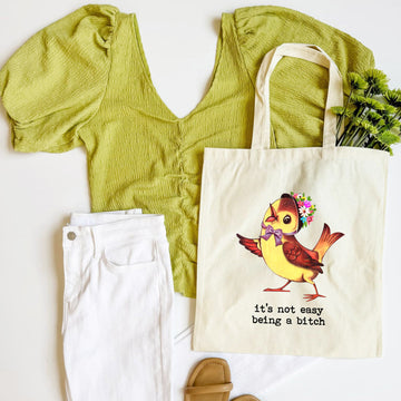 Green top, white pants, beige sandals, and a tote bag with a colorful bird illustration on a white background