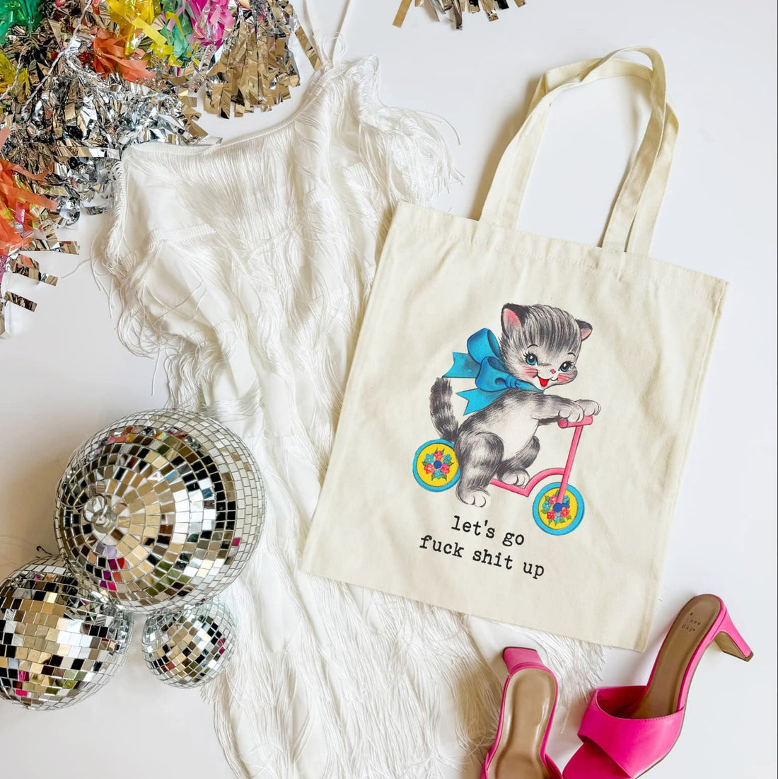 Tote bag with a cat illustration and text, surrounded by disco balls and high heels on a white surface.