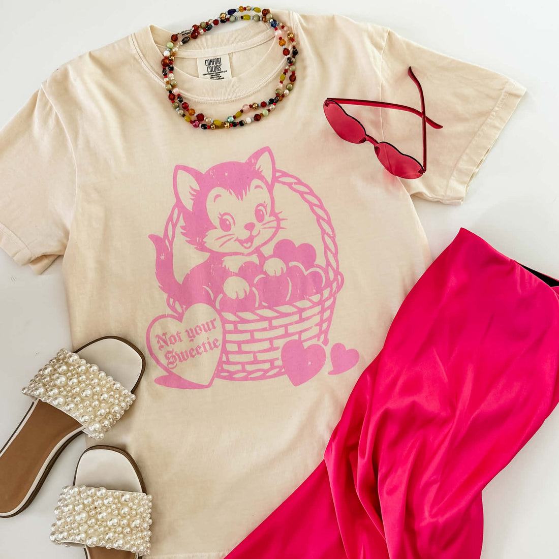Beige t-shirt with pink cat graphic, sunglasses, pink scarf, and pearl sandals on a light background