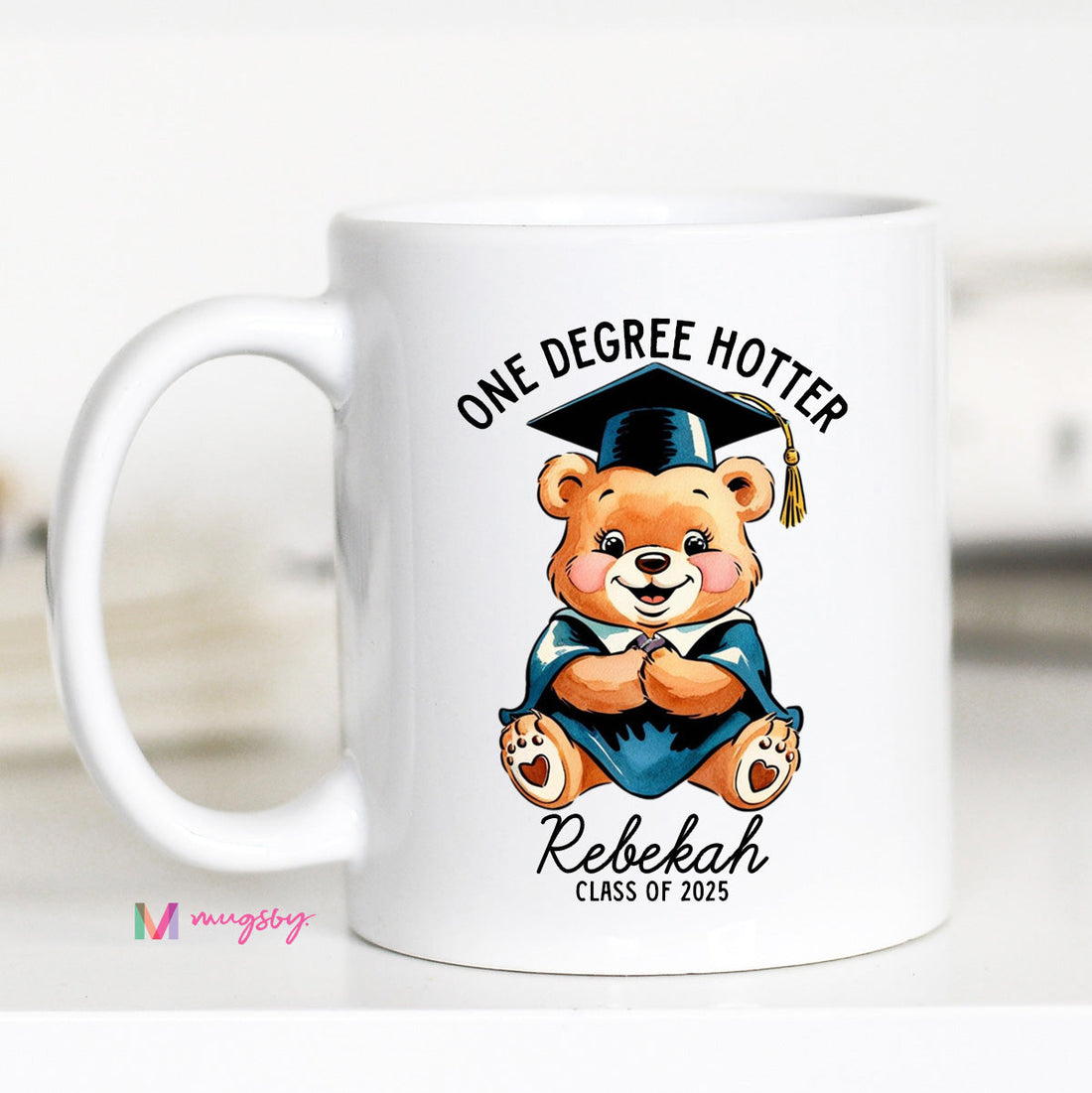 A white ceramic coffee mug with a black handle featuring a cartoon bear wearing a graduation cap and the text &