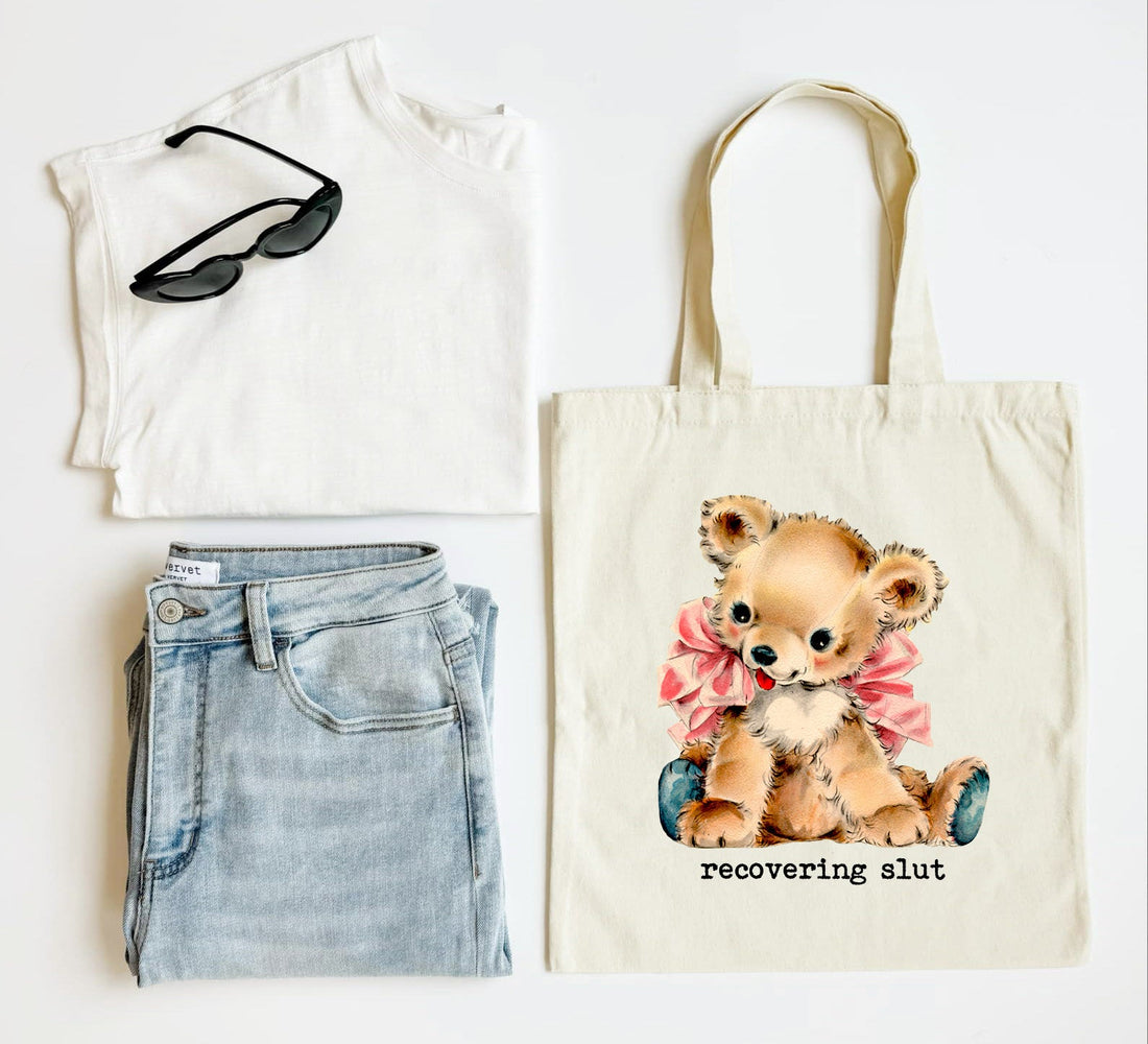 Tote bag with teddy bear design and text, paired with a white top, jeans, and sunglasses on a light background.