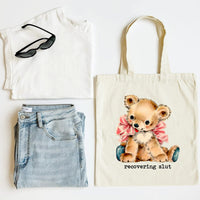 Tote bag with teddy bear design and text, paired with a white top, jeans, and sunglasses on a light background.