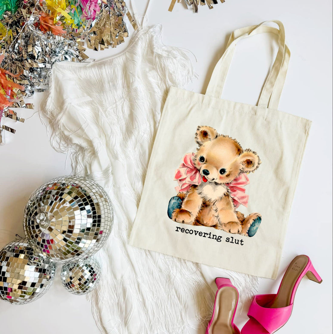 Tote bag with teddy bear graphic and text, surrounded by disco balls and high heels on a white surface.